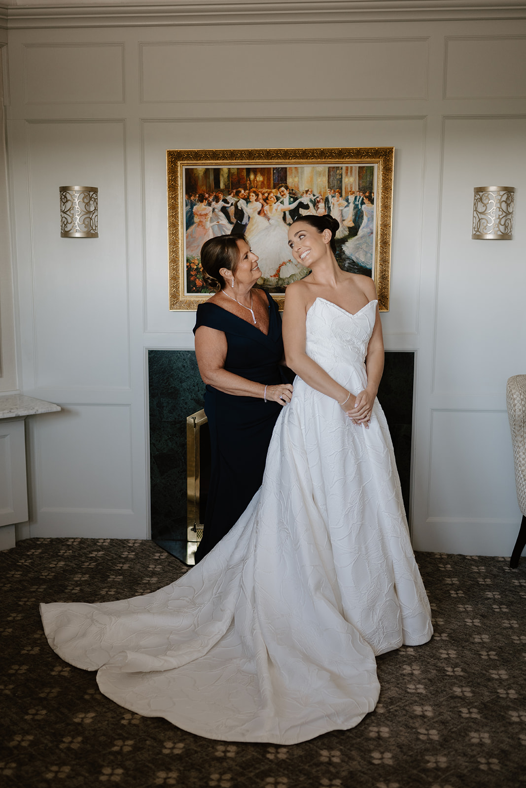 Bride and her mother smiling warmly at each other while posing in front of a fireplace.